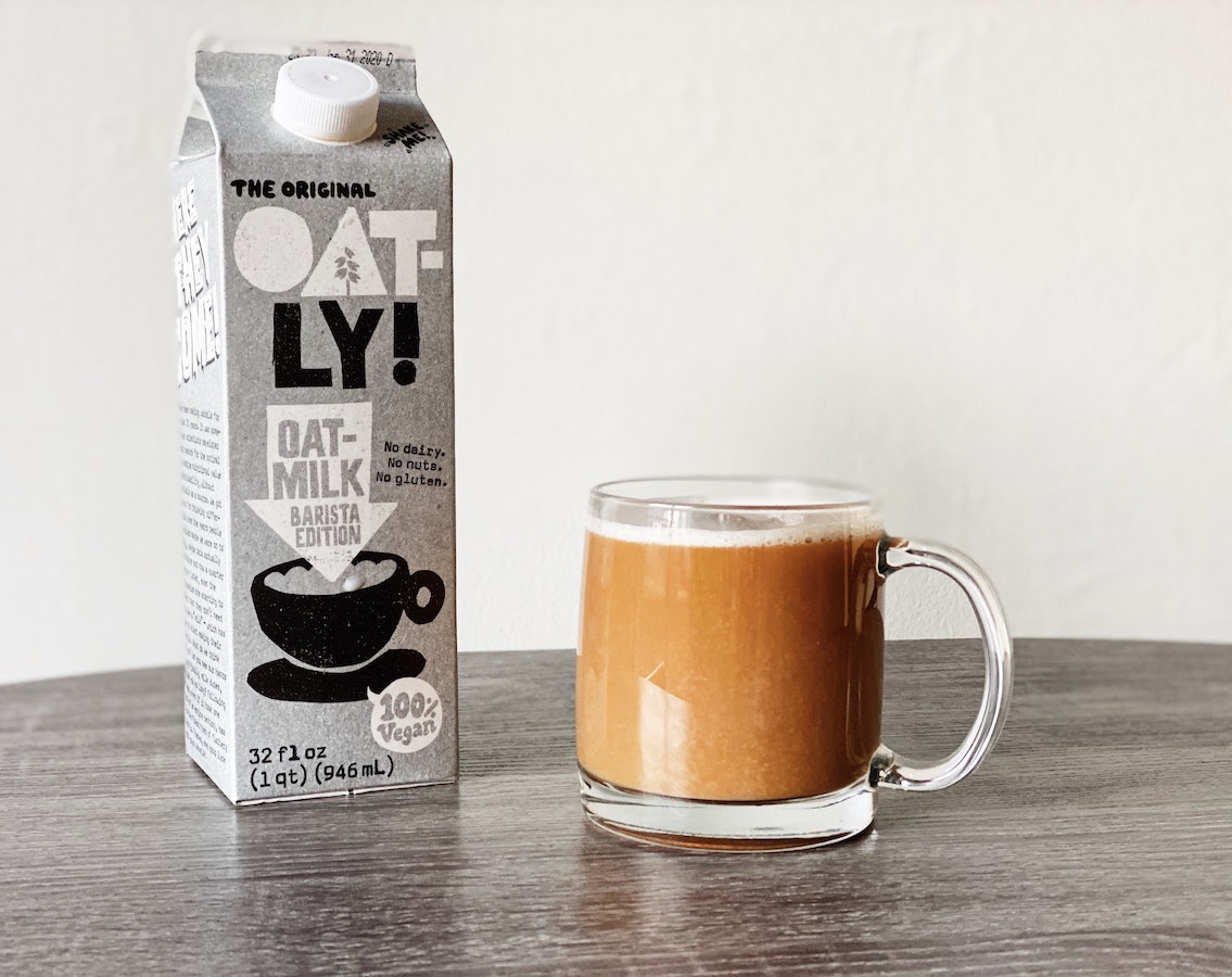 On a grey table is a cup of coffee with milk in it. Next to the cup of coffee is a cartoon of oatly oat milk.