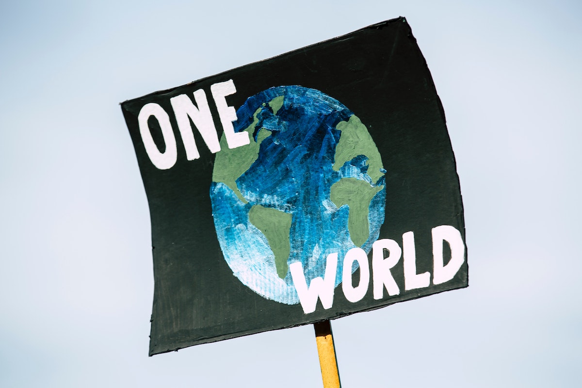 A paper sign being held up on a wooden stick. The sign is black with a drawing of the planet on it. There is writing that says 'one world'