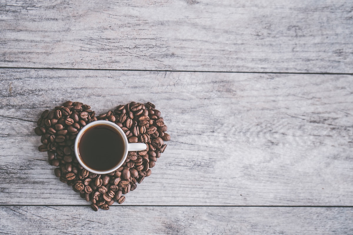 A wooden background that looks like a table top. To the left of the photo is coffee beans shaped into a heart. In the centre of heart is a cup of black coffee.