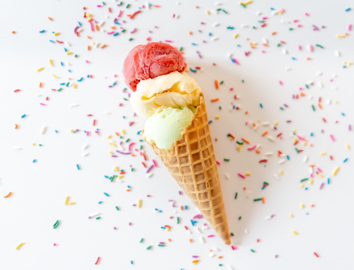 On a white background is an ice cream cone with three scoops of ice cream. The scoops are different colours, one is light green, another yellow, and one is pink. There are coloured sprinkles surrounding the cone.