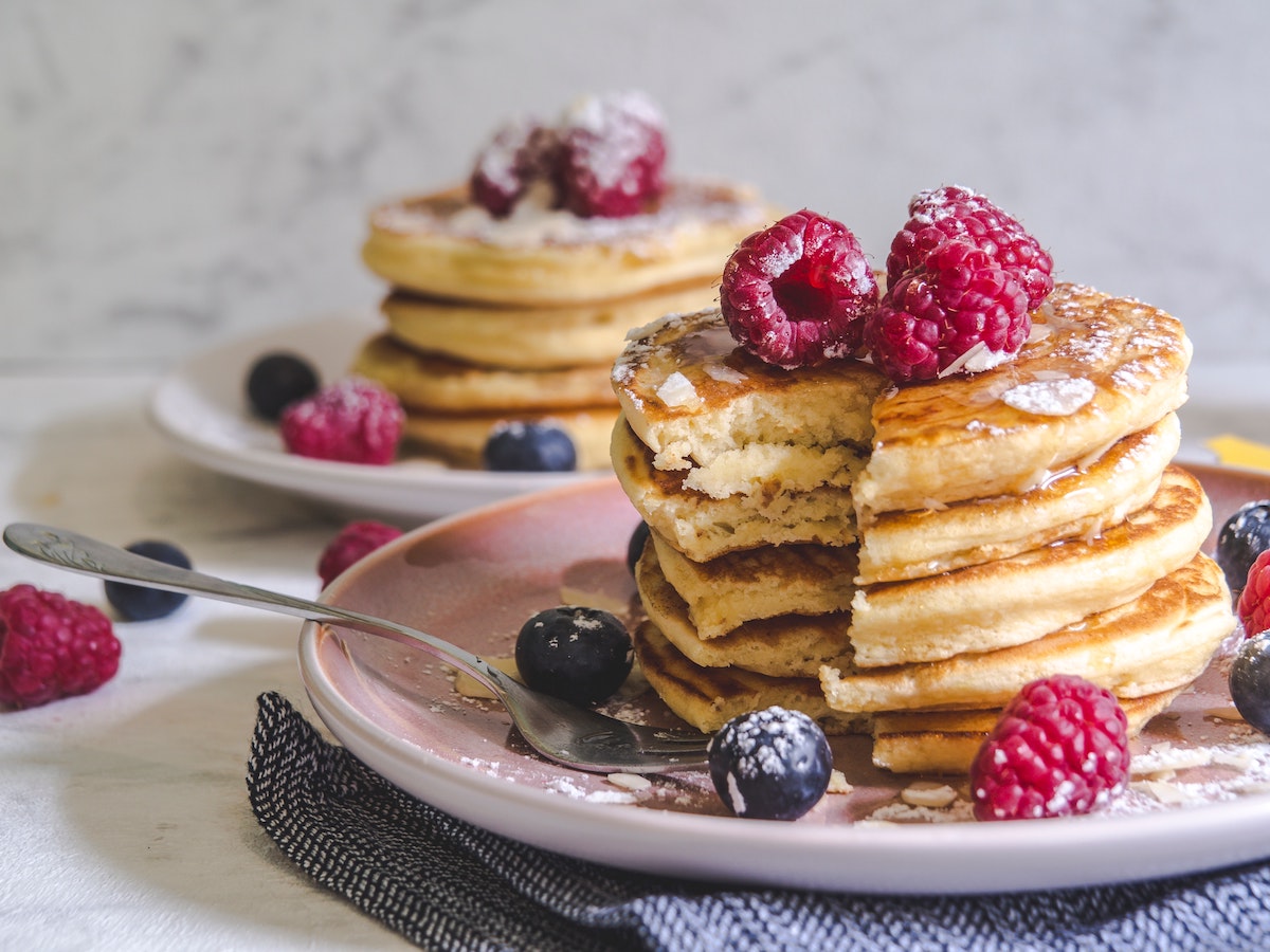 A stack of American pancakes on a pink plate. Pancakes are covered in icing sugar and berries.