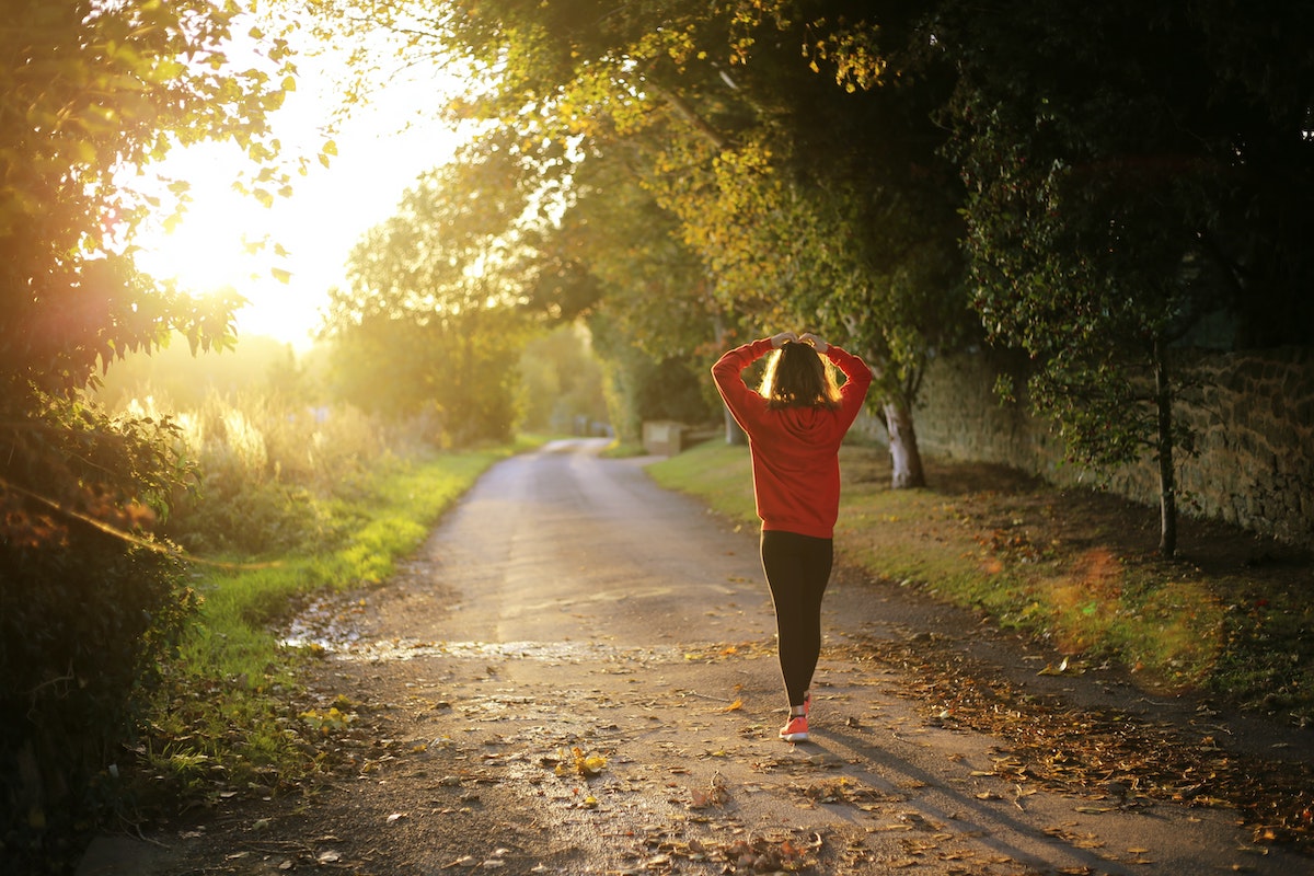woman in an orange jumper and black leggings facing away from the camera. She is walking down a tree lined road with the sunset ahead of her.
